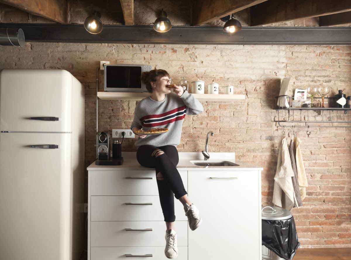 girl sitting on counter in kitchenette eating pizza next to a fridge and sink