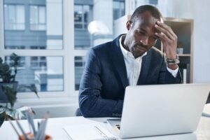 man sitting at desk looking at computer and holding his head