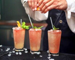 hands squeezing lemon over three alcoholic drinks on bartop