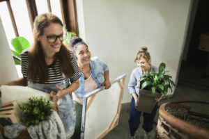 three women walking up the stairs to their new apartment holding plants and pictures