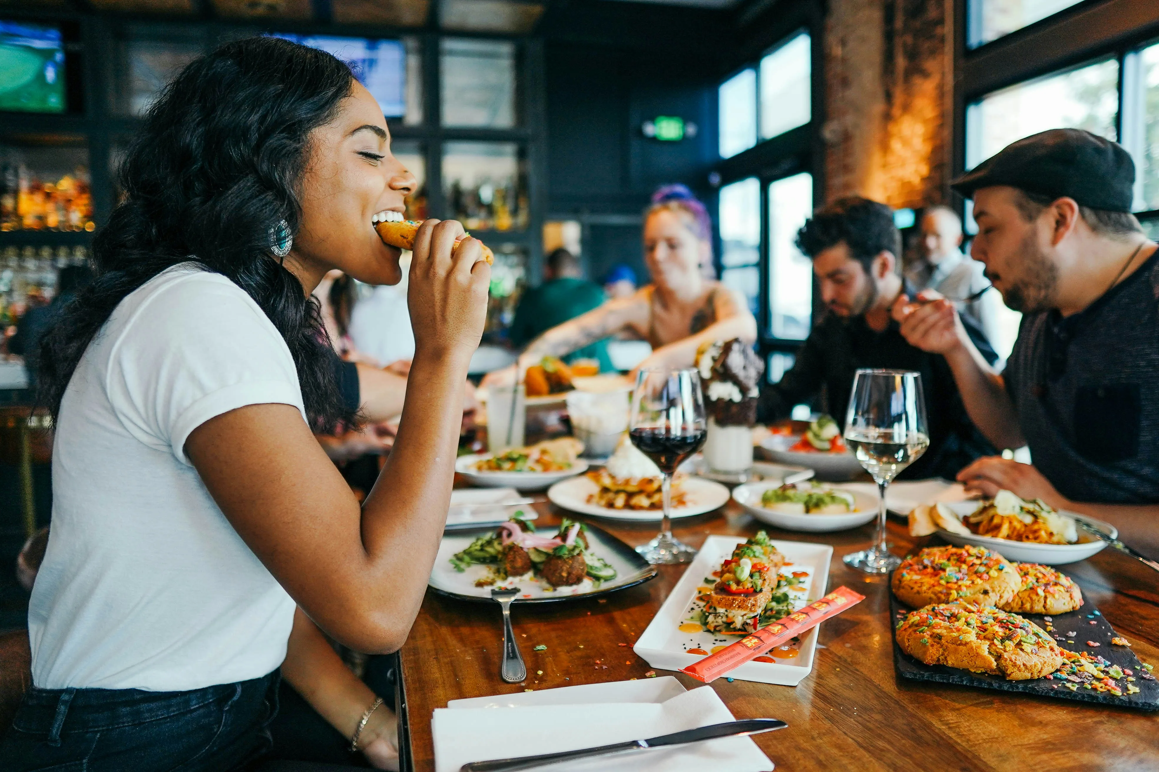 a single woman taking a bite of food surrounded by friends at a restaurant