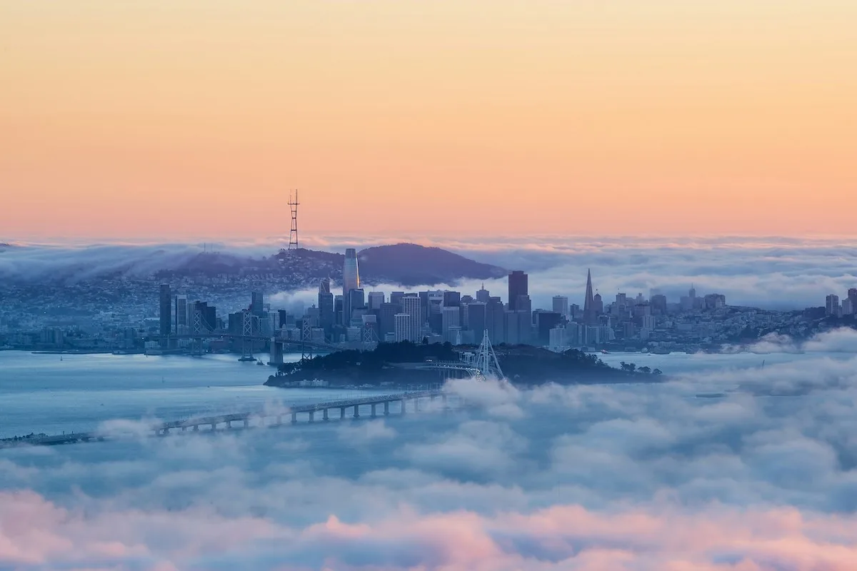 San Francisco skyline emerging through fog at sunrise with the Bay Bridge in view โ highlighting the cityโs mix of beauty and high cost of living that renters weigh in 2025.